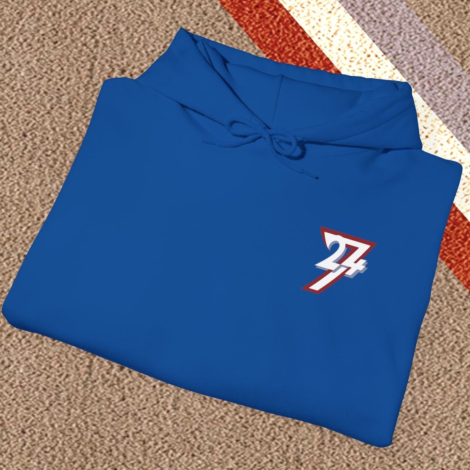 Blue stylish hoodie with twentyfourseven logo, folded on textured carpet with red, white, and purple stripes.