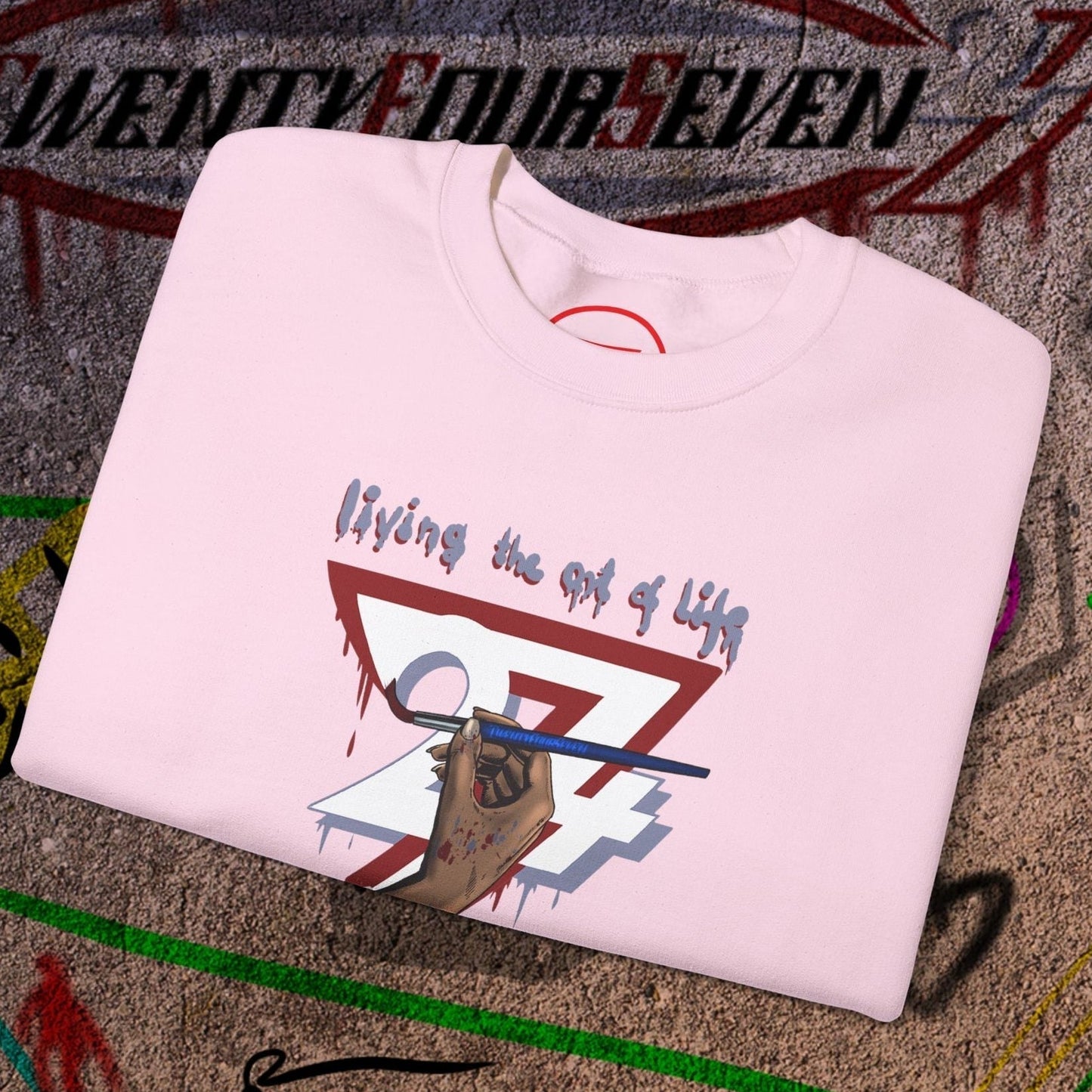 Folded light pink artist brush sweatshirt with hand-drawn bohemian design and "living the art of life" text on concrete background
