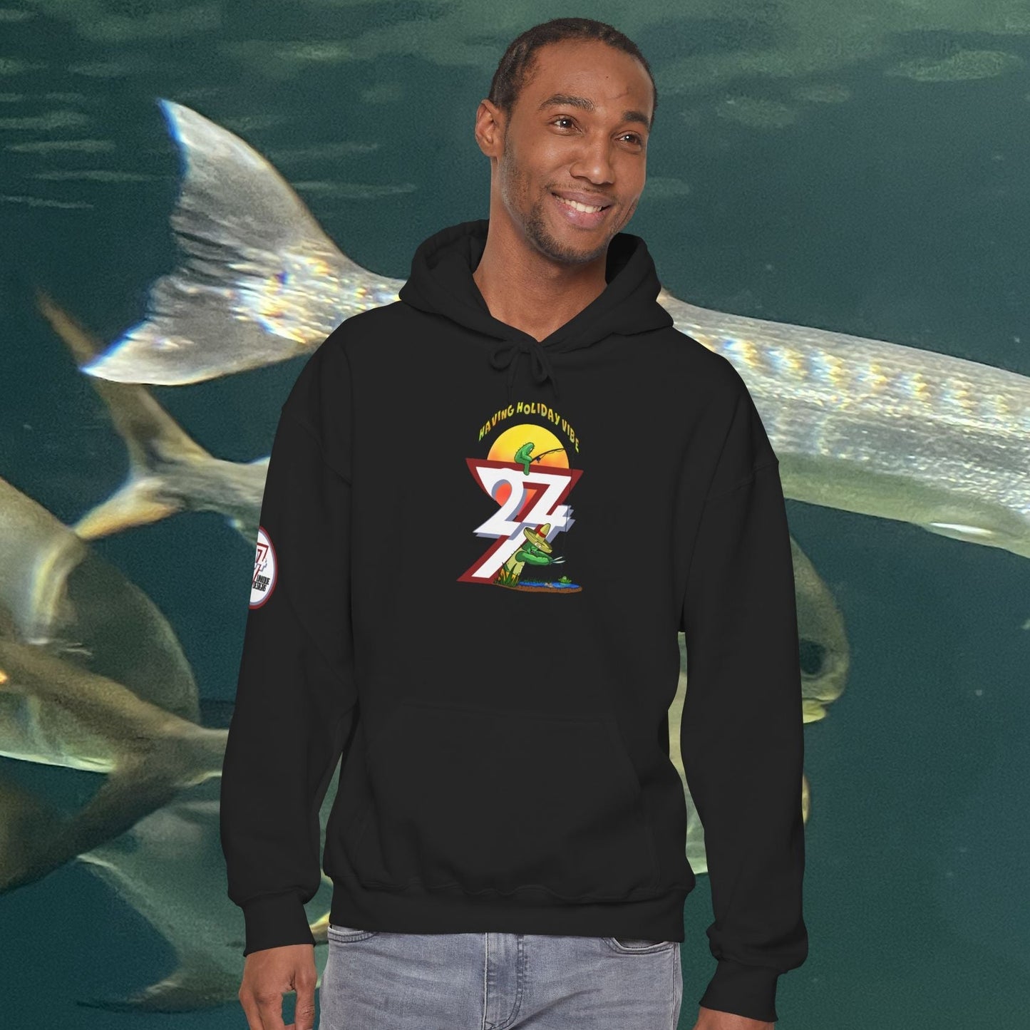 Man wearing black Funny Fishing Hoodie with colorful cactus and fishing design, standing in front of underwater fish background
