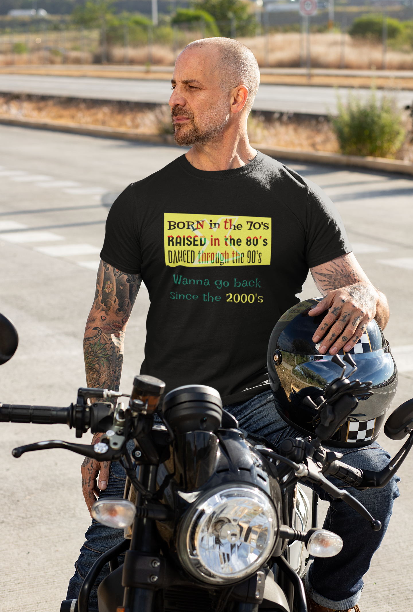 Man wearing black Gen-X tee with retro print sitting on motorcycle holding helmet outdoors