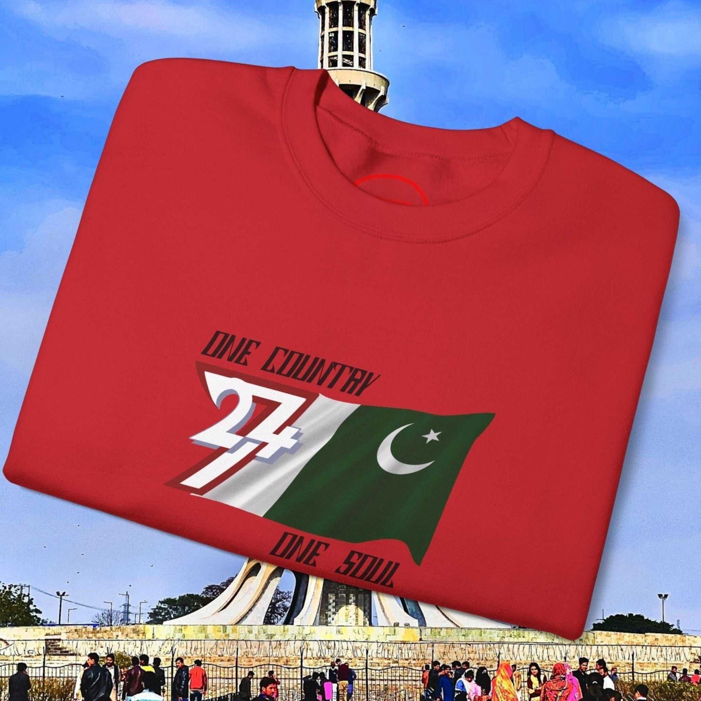 Pakistan Flag Sweatshirt red crewneck with hand-drawn Pakistan flag and "One Country One Soul" slogan, folded against Minar-e-Pakistan background.