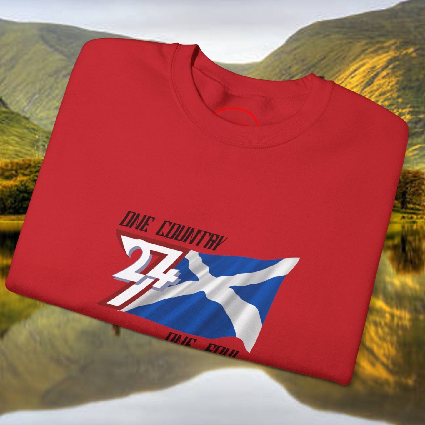 Red Scotland Saltire crewneck sweatshirt with hand-drawn St. Andrew’s Cross design folded on scenic Scottish landscape background