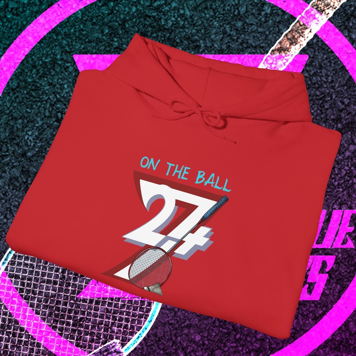 Red badminton hoodie with "On the Ball 24/7" graphic design, folded with drawstring hood and kangaroo pocket, placed on a badminton court background