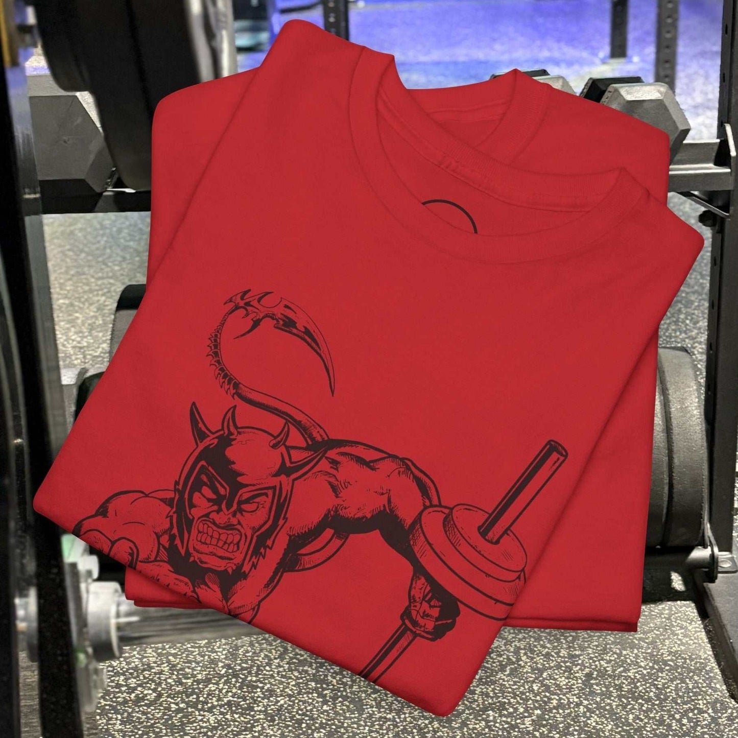 Red gym tee with hand-drawn comic-style fantasy artwork of a muscular devil holding a barbell, folded on gym floor equipment