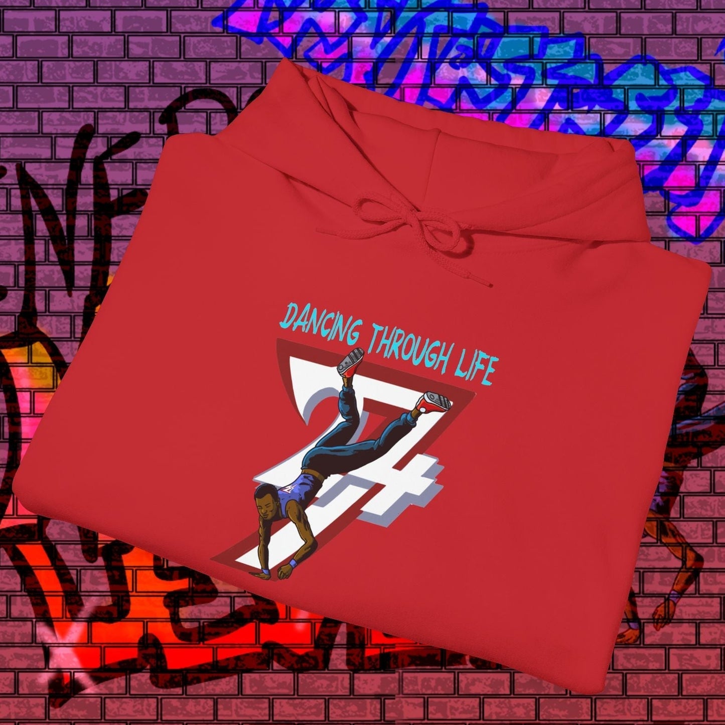 Red street dance hoodie with hand-drawn dancer illustration and "Dancing Through Life" text on front, folded on graffiti brick wall background