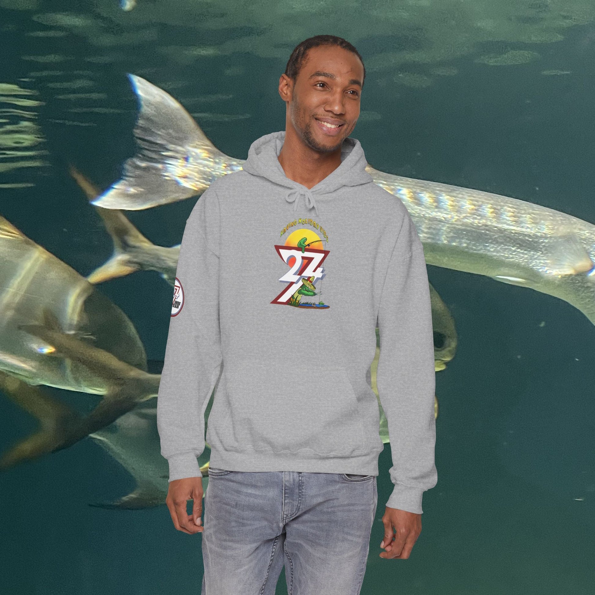 man wearing grey funny fishing hoodie with cactus design smiling by aquarium tank with fish swimming behind