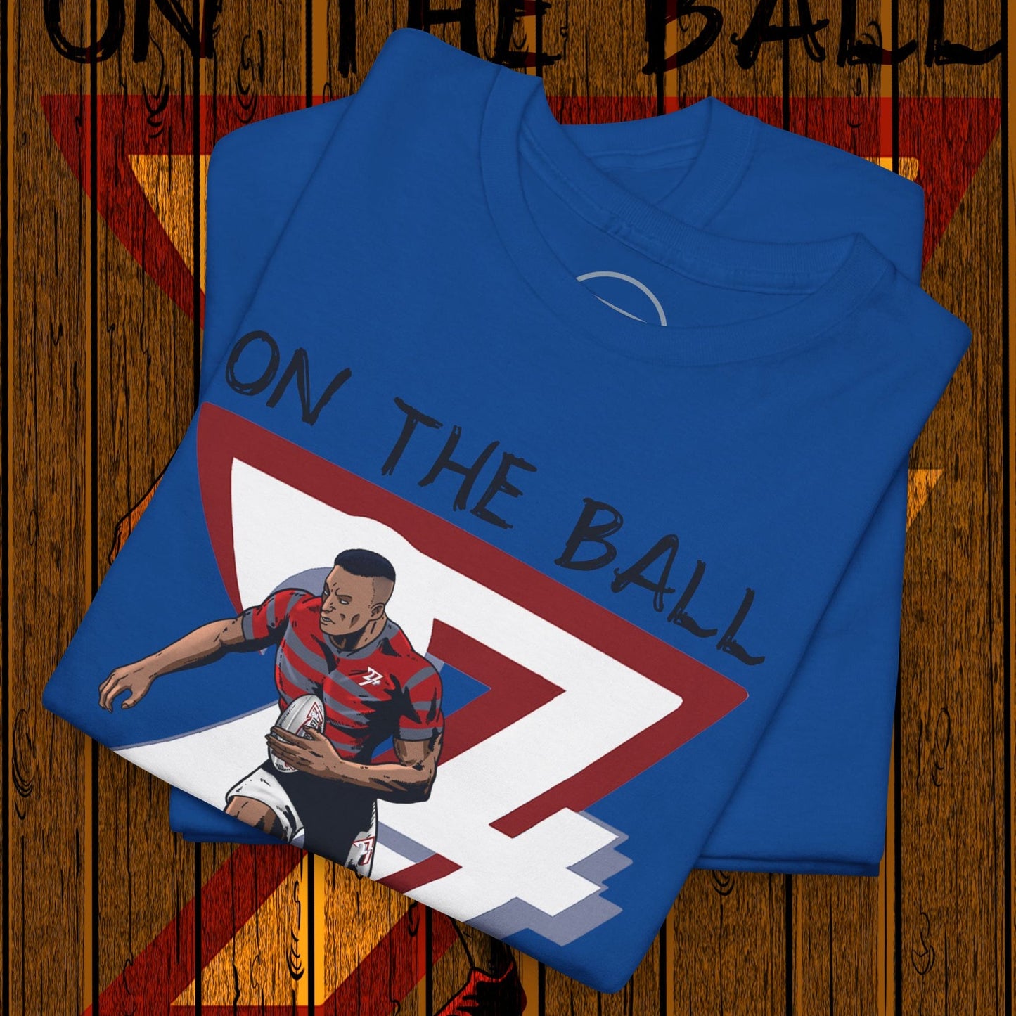 Blue rugby sports tee with hand-drawn player illustration and "On The Ball" text, folded on wooden floor background