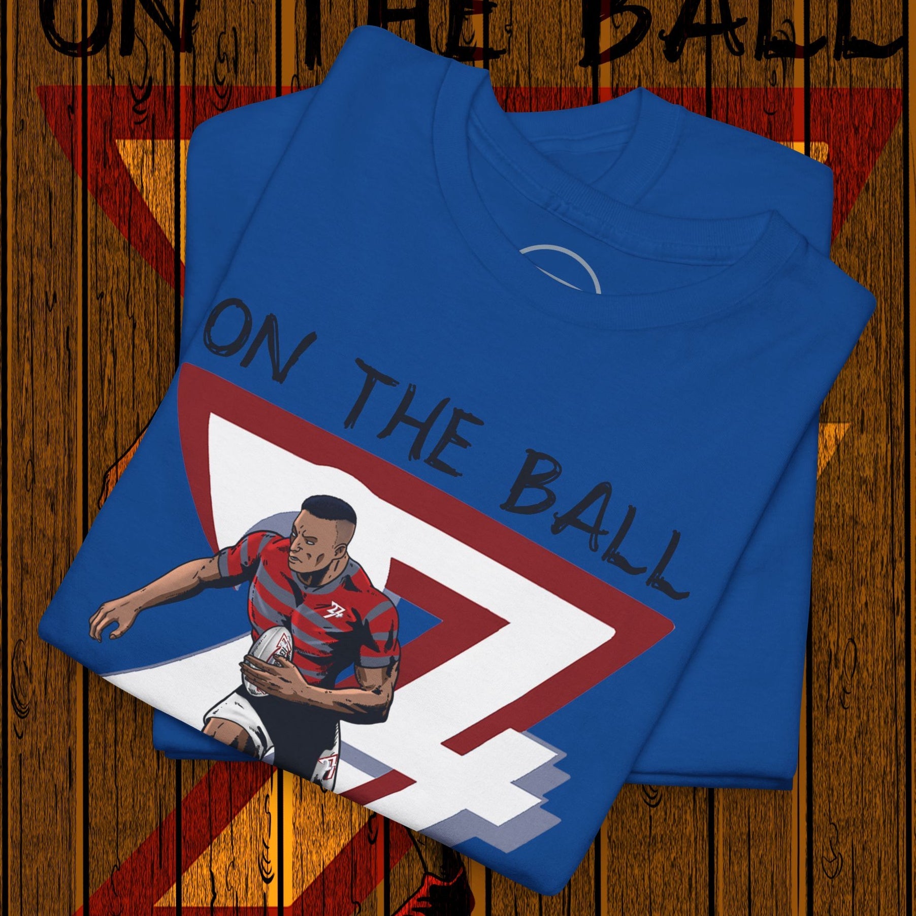Blue rugby sports tee with hand-drawn player illustration and "On The Ball" text, folded on wooden floor background