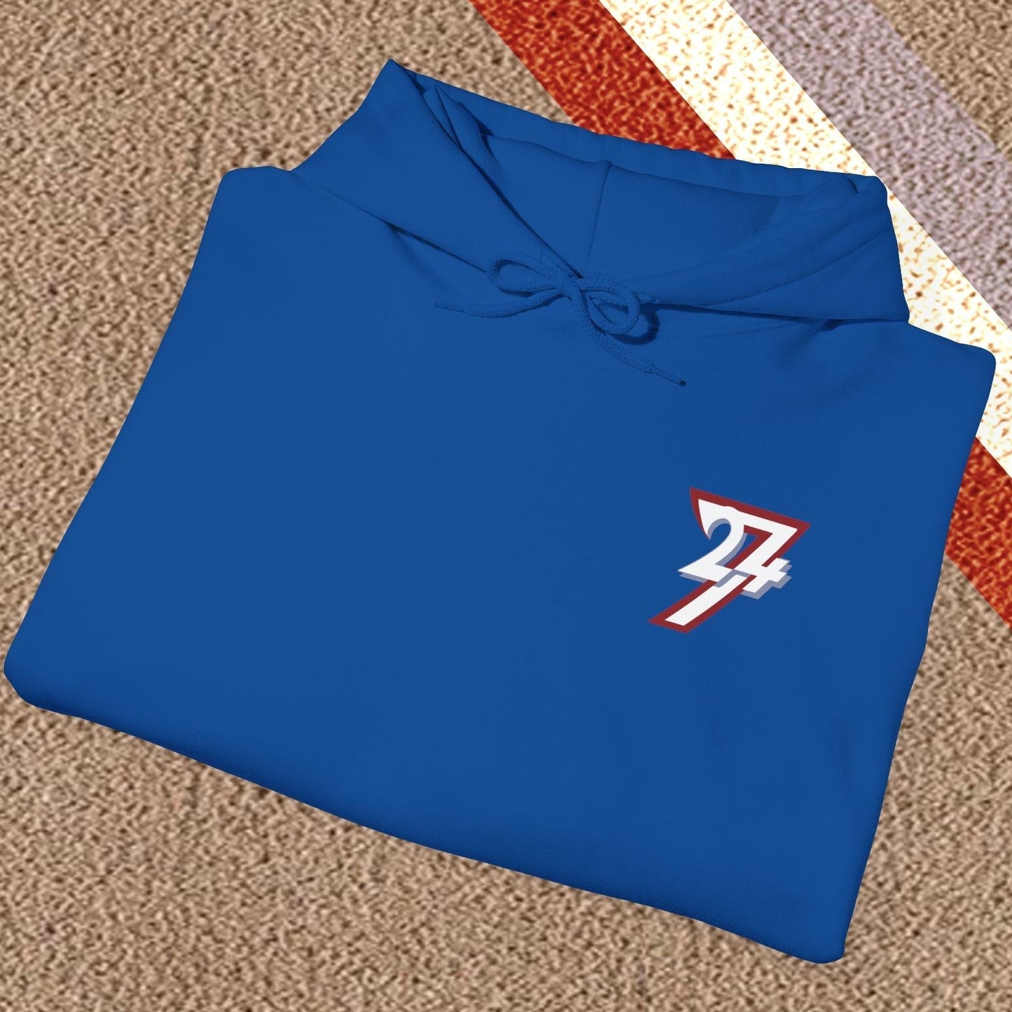 Blue stylish hoodie with twentyfourseven logo, folded on textured carpet with red, white, and purple stripes.