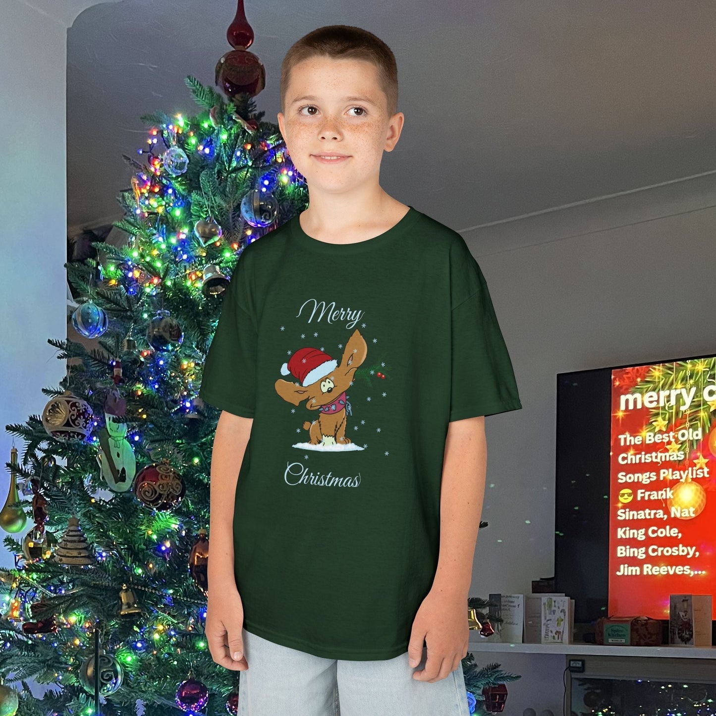 Child wearing green kids Christmas tee with festive puppy design and "Merry Christmas" text by decorated Christmas tree
