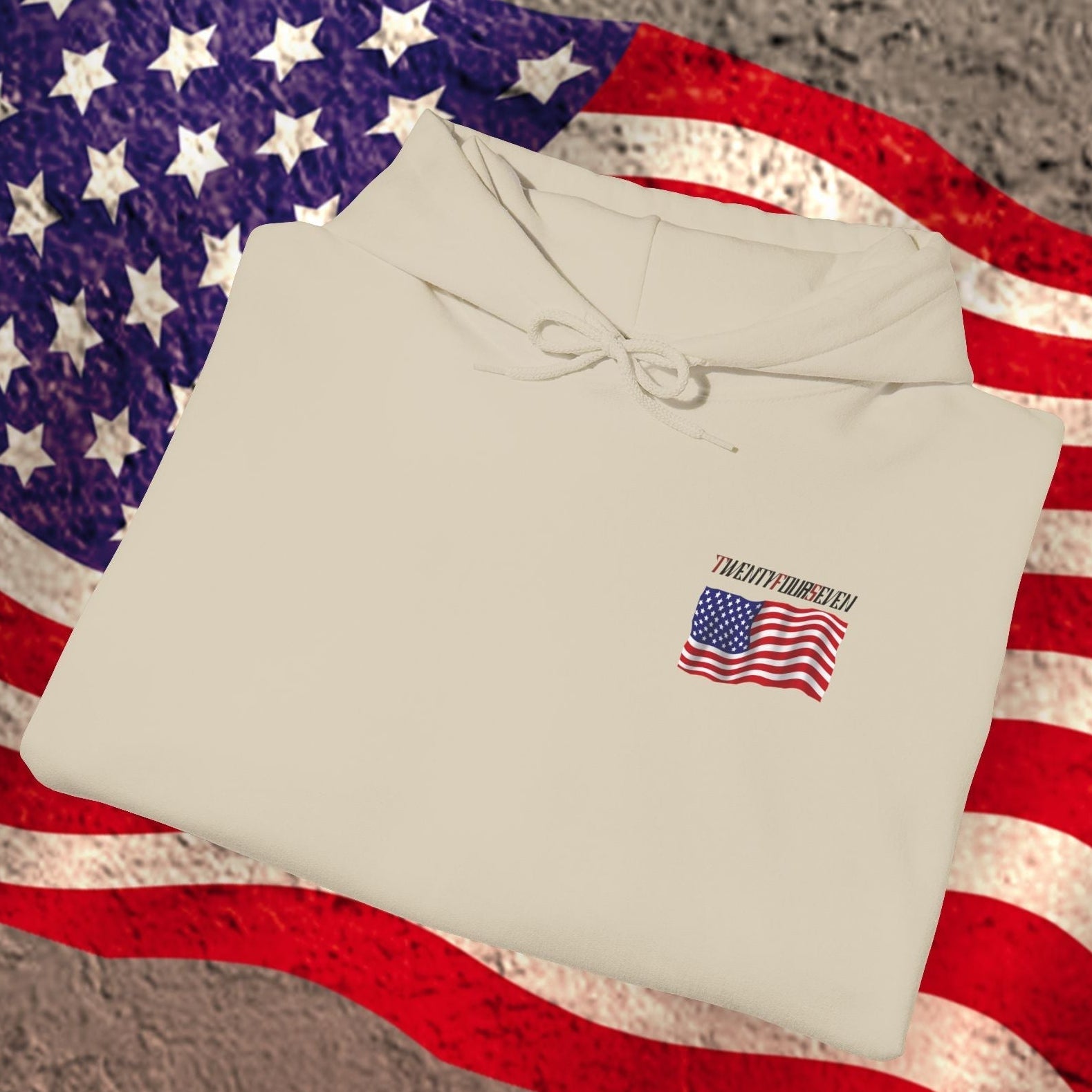 Folded beige American flag hoodie with small US flag print on chest, placed on large American flag background.