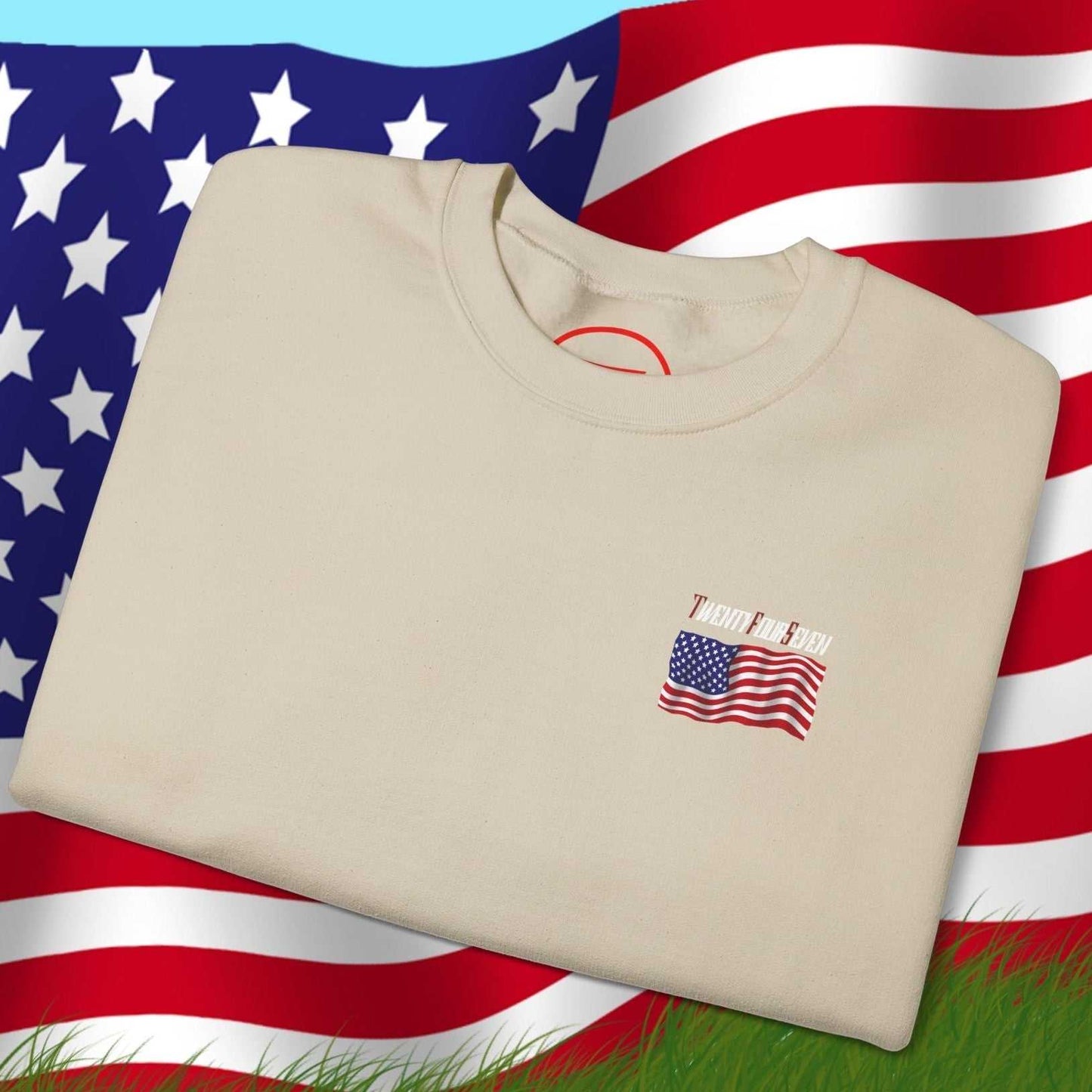 Folded beige US Flag Sweatshirt with small American flag design on chest, placed on grass with large waving US flag background