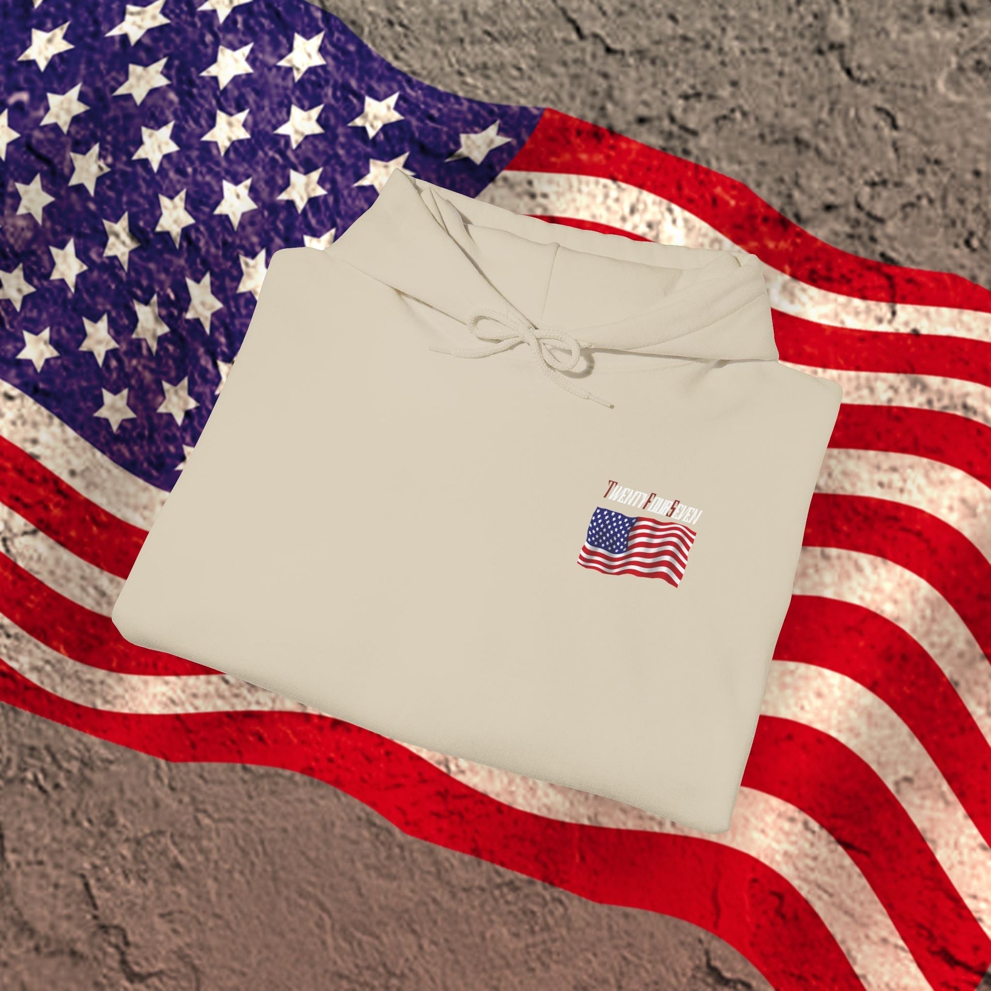 Folded beige US flag hoodie with small American flag emblem on chest, placed on a large American flag background