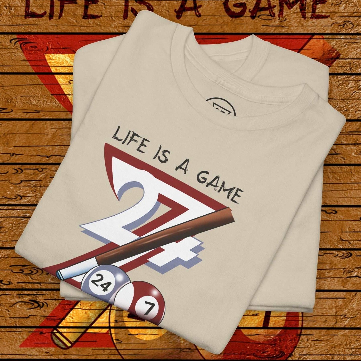 Folded beige pool tee with "Life is a Game" text and billiards design featuring cue and balls on wooden textured background
