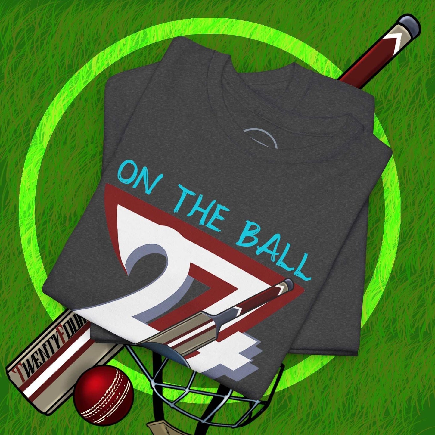 Folded dark gray cricket tee with hand-drawn "ON THE BALL 24/7" design, cricket bat, ball, and helmet on grass background