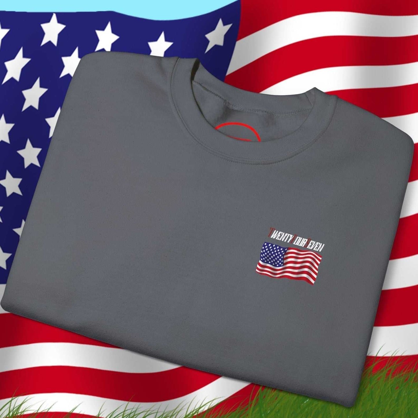 Folded gray US flag sweatshirt with hand-drawn American flag design on chest, displayed on large American flag background outdoors
