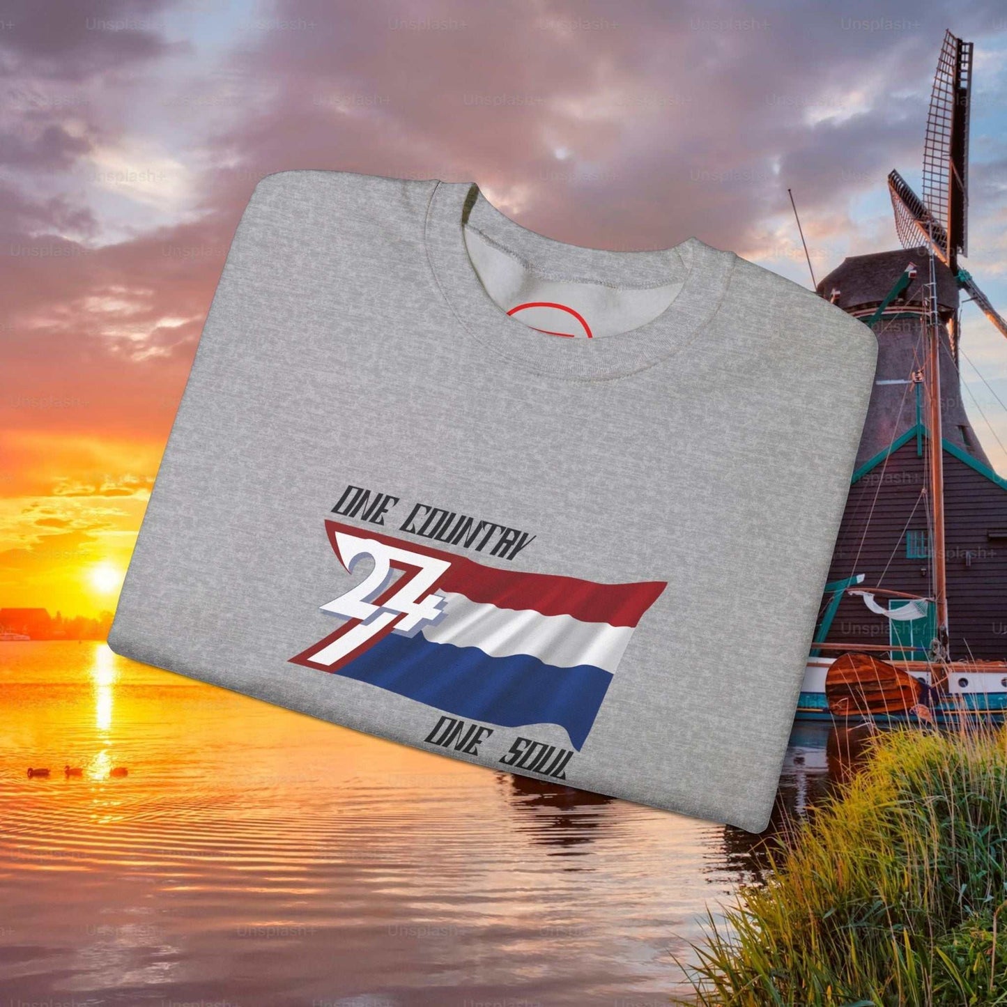 Folded grey Netherlands flag sweatshirt with red, white, and blue design and "One Country One Soul" text on scenic sunset background
