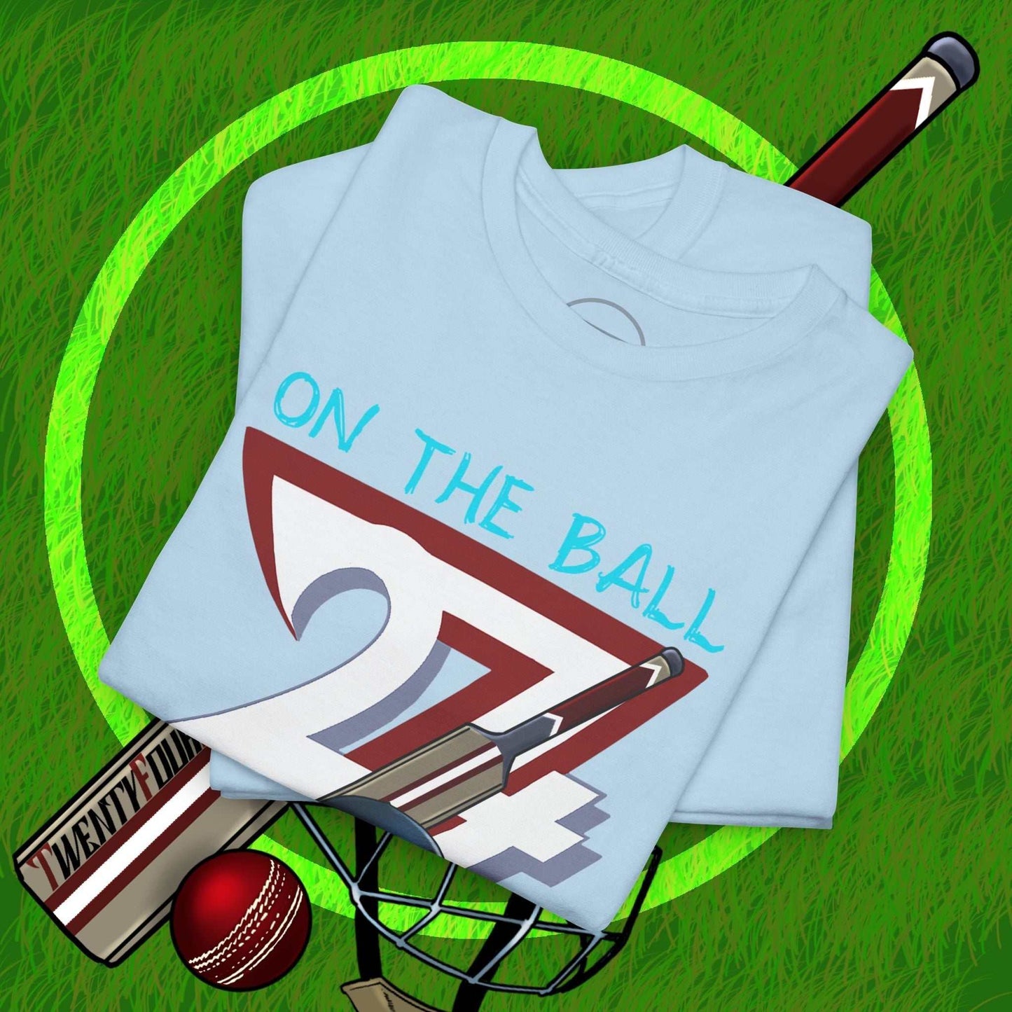 Folded light blue cricket tee with "On The Ball 24/7" graphic, cricket bat, ball, and helmet on green grass background