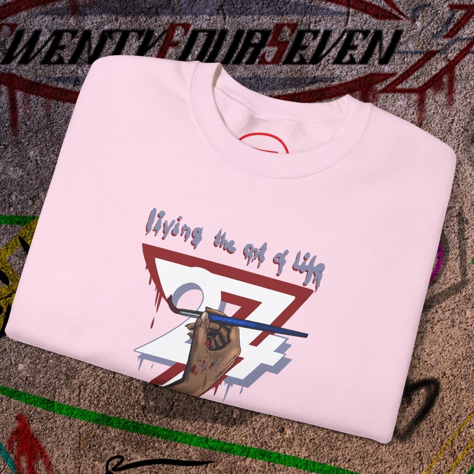 Folded light pink artist brush sweatshirt with hand-drawn bohemian design and "living the art of life" text on concrete background