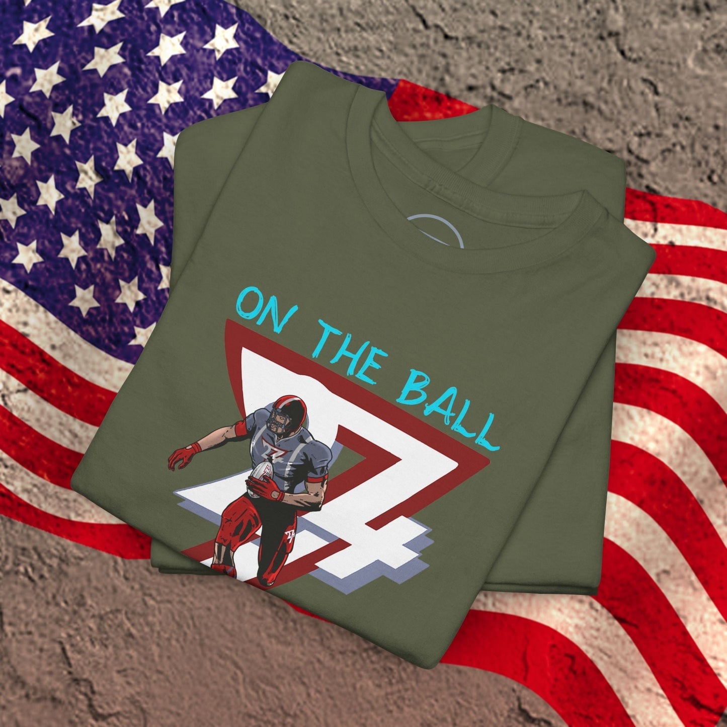Folded olive green American football tee with comic-style player graphic and "ON THE BALL" text on chest over American flag background
