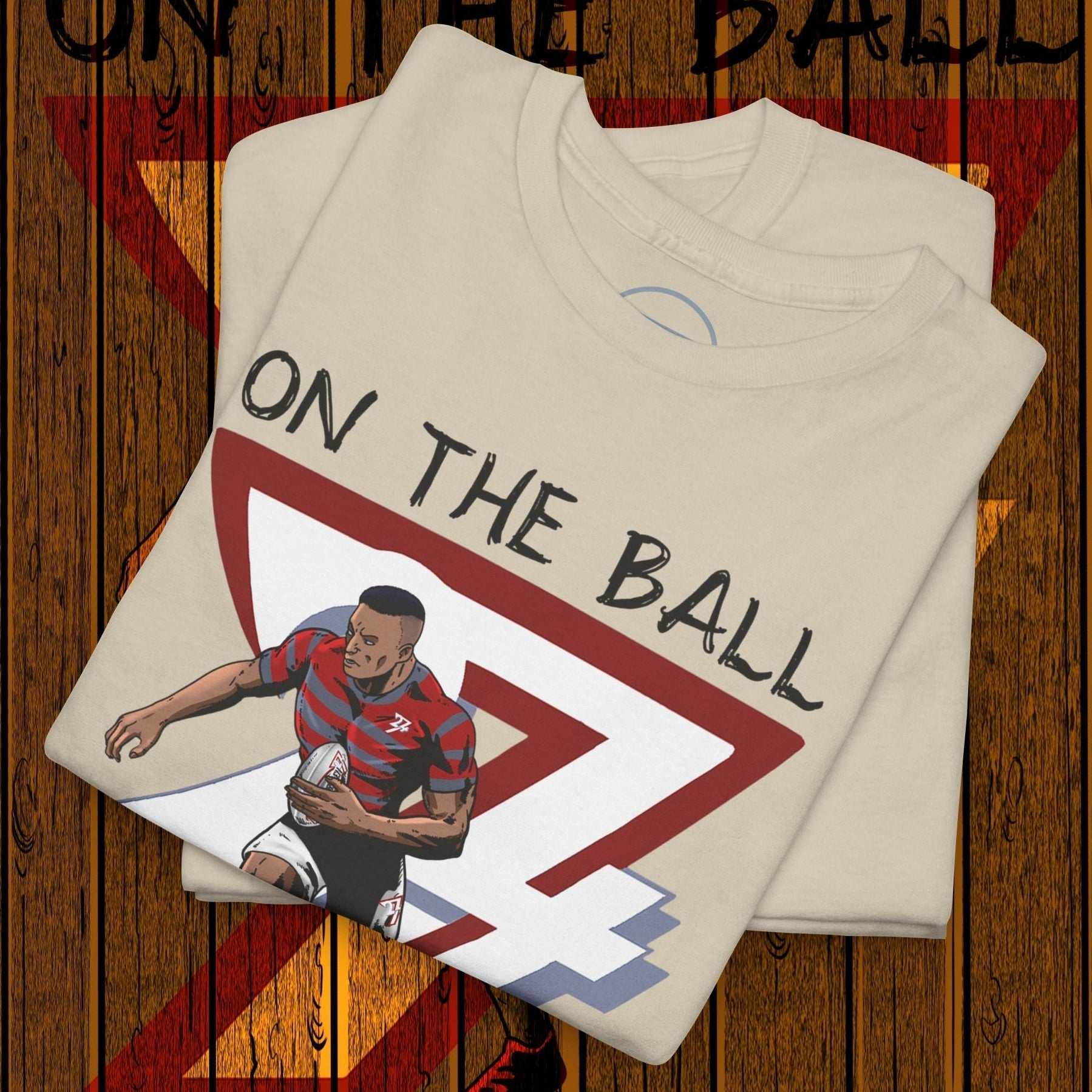 Folded rugby sports tee with hand-drawn rugby player illustration and "On The Ball" text on wooden background