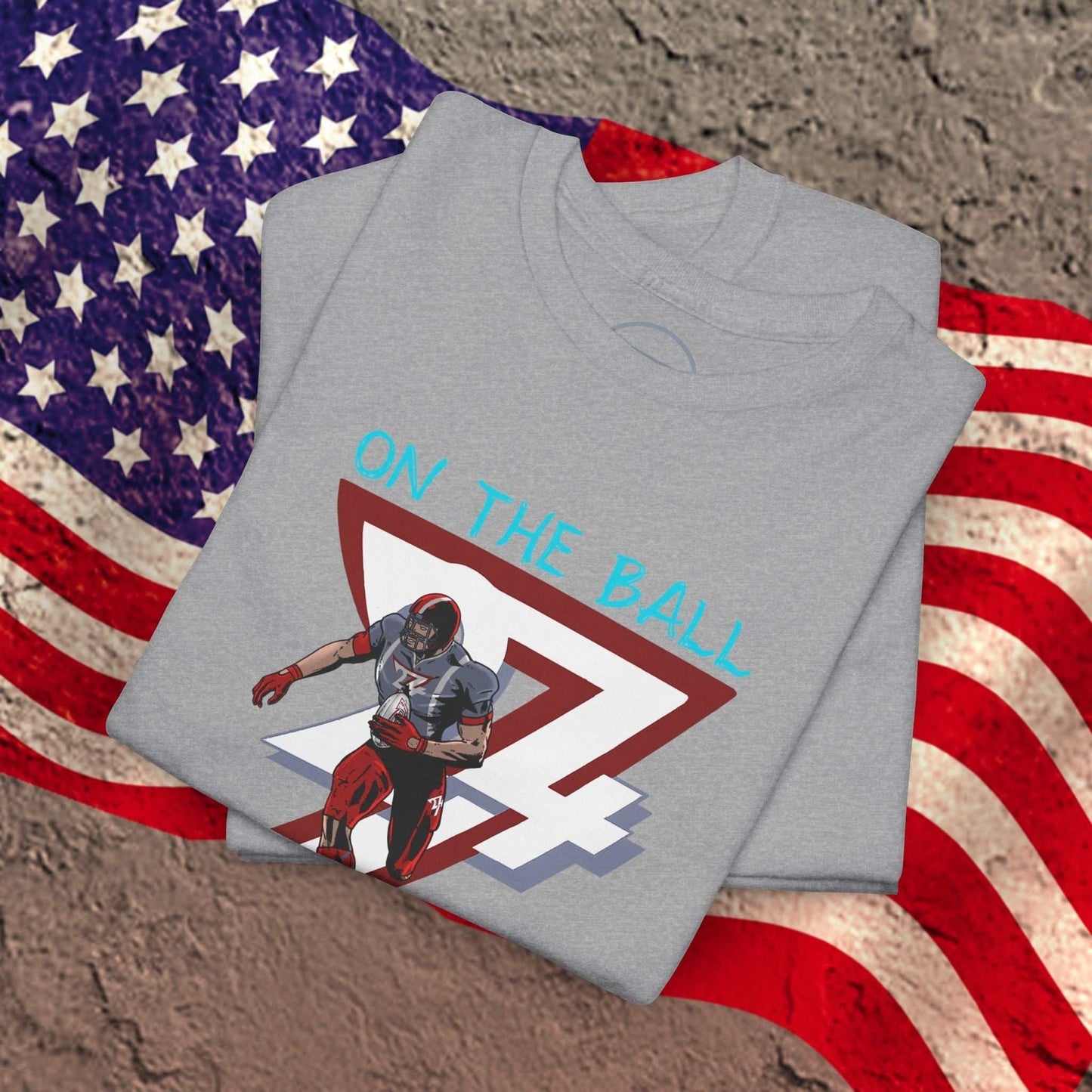 Gray American Football Tee with hand-drawn comic-style player graphic and "On The Ball" text, folded on an American flag background