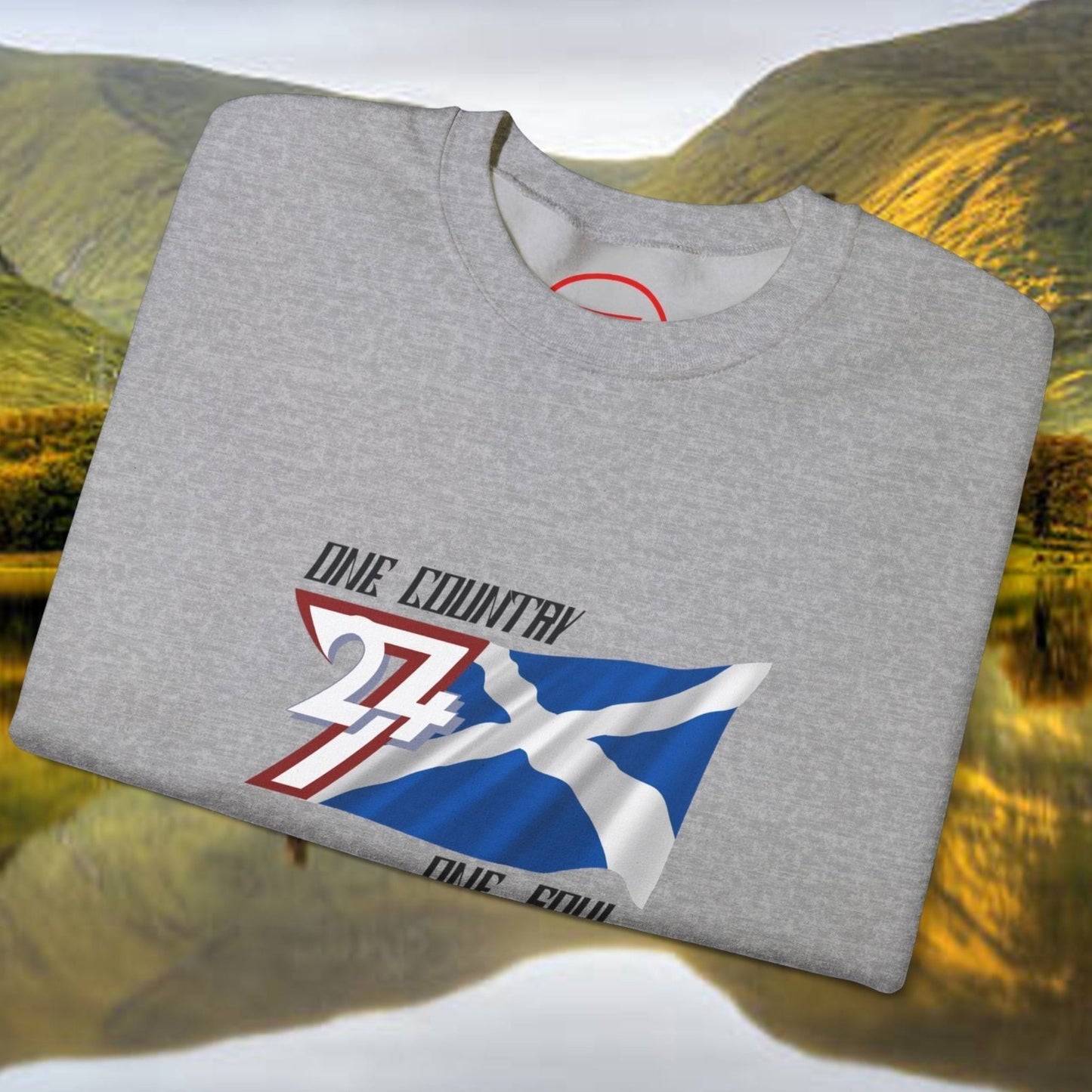 Gray Scotland Saltire crewneck sweatshirt with hand-drawn St. Andrew’s Cross design folded on a scenic Scottish landscape background