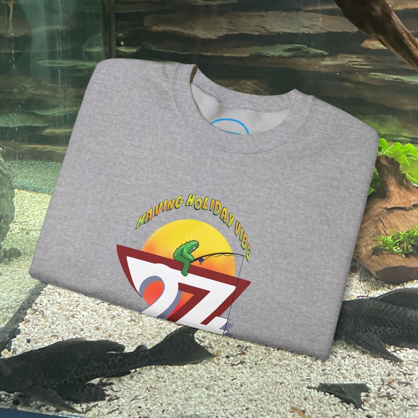 Gray fishing cactus sweatshirt with colorful cartoon design and "Having Holiday Vibes" text folded on aquarium background