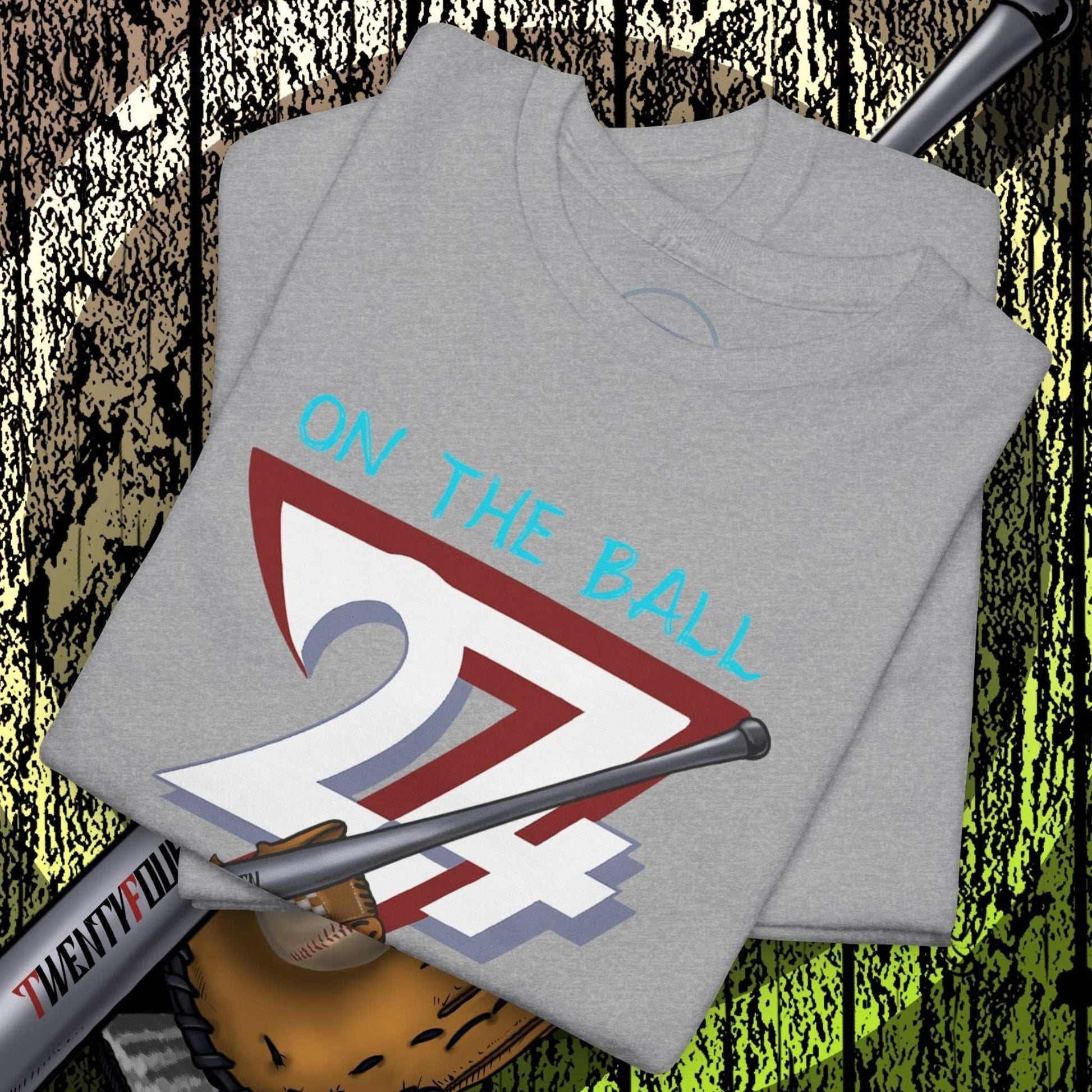 Grey Baseball Illustrated Tee with hand-drawn "On The Ball 24/7" design, paired with baseball bat and glove on wooden background