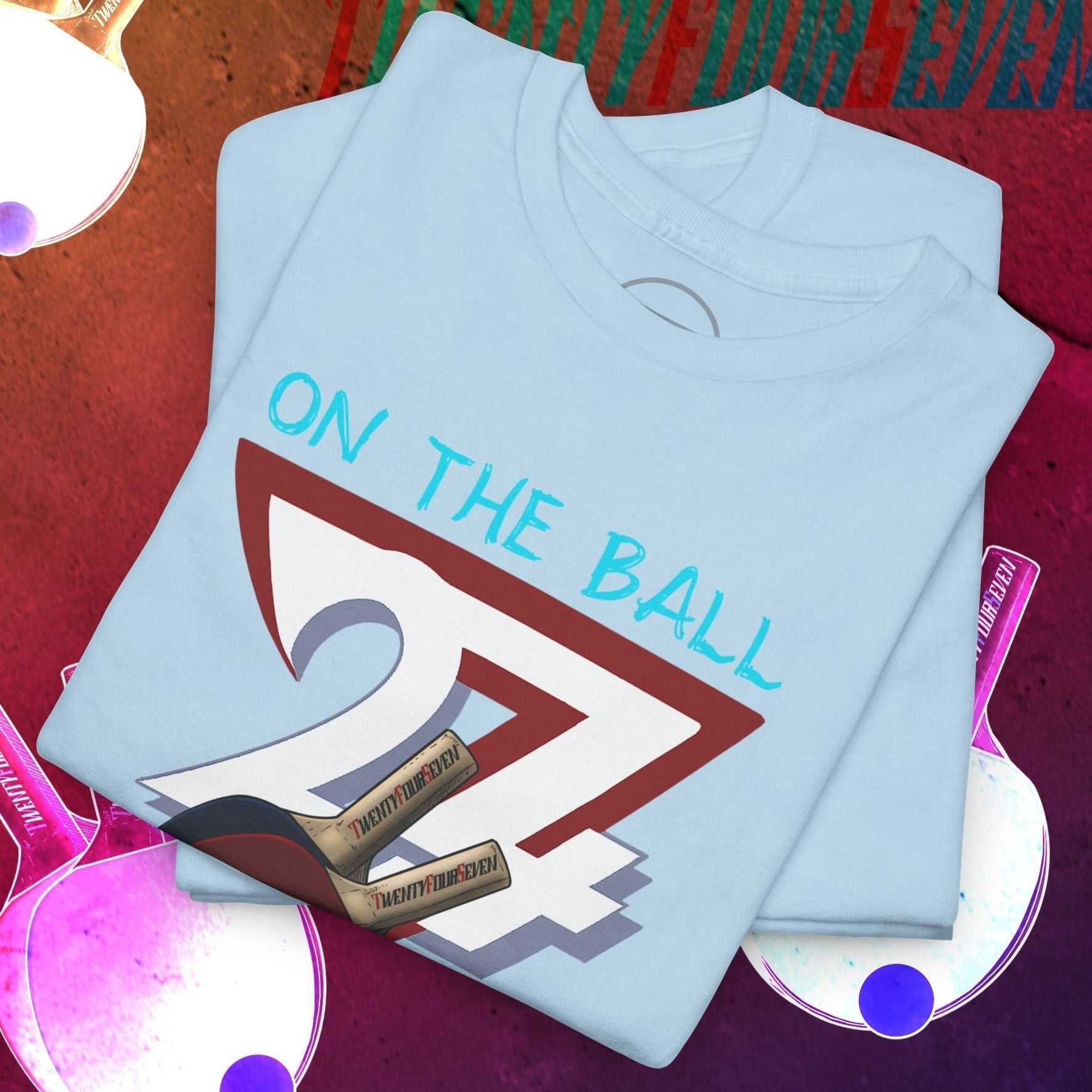 Light blue Ping-Pong T-shirt featuring a hand-drawn "On The Ball 24/7" sports illustration with folded design on a red and purple background with ping-pong paddles and balls