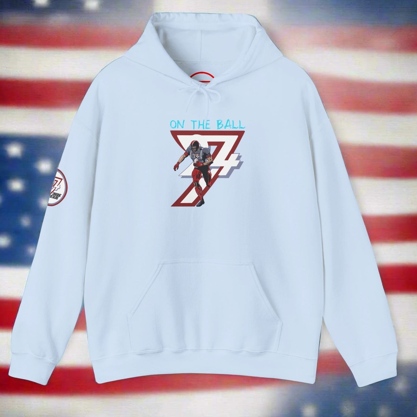 Light blue american football hoodie with hand-drawn player design and "On The Ball" text on front, kangaroo pocket, and adjustable hood against blurred American flag background