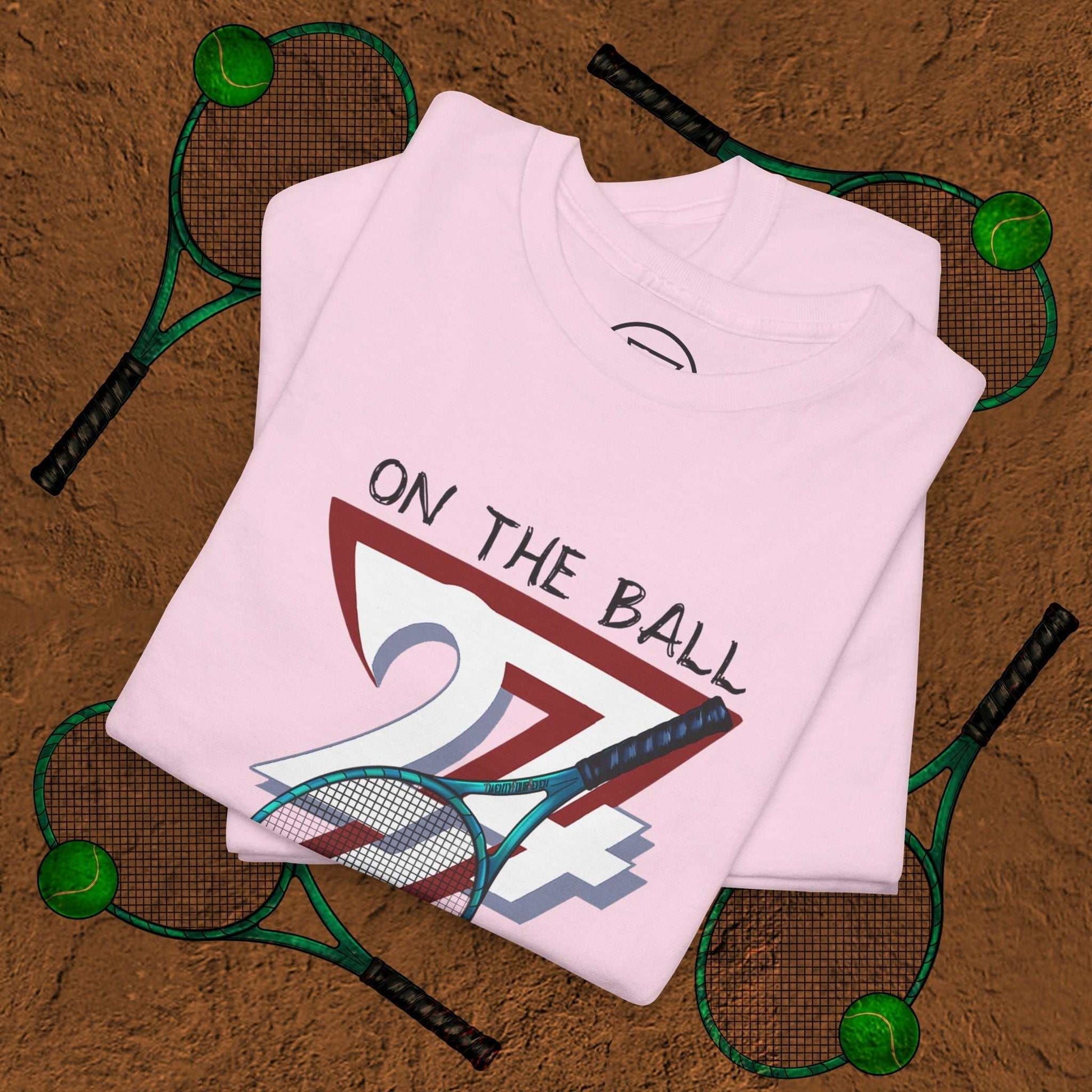 Pink tennis tee with "On the Ball 24/7" hand-drawn design and tennis racquets on clay court background