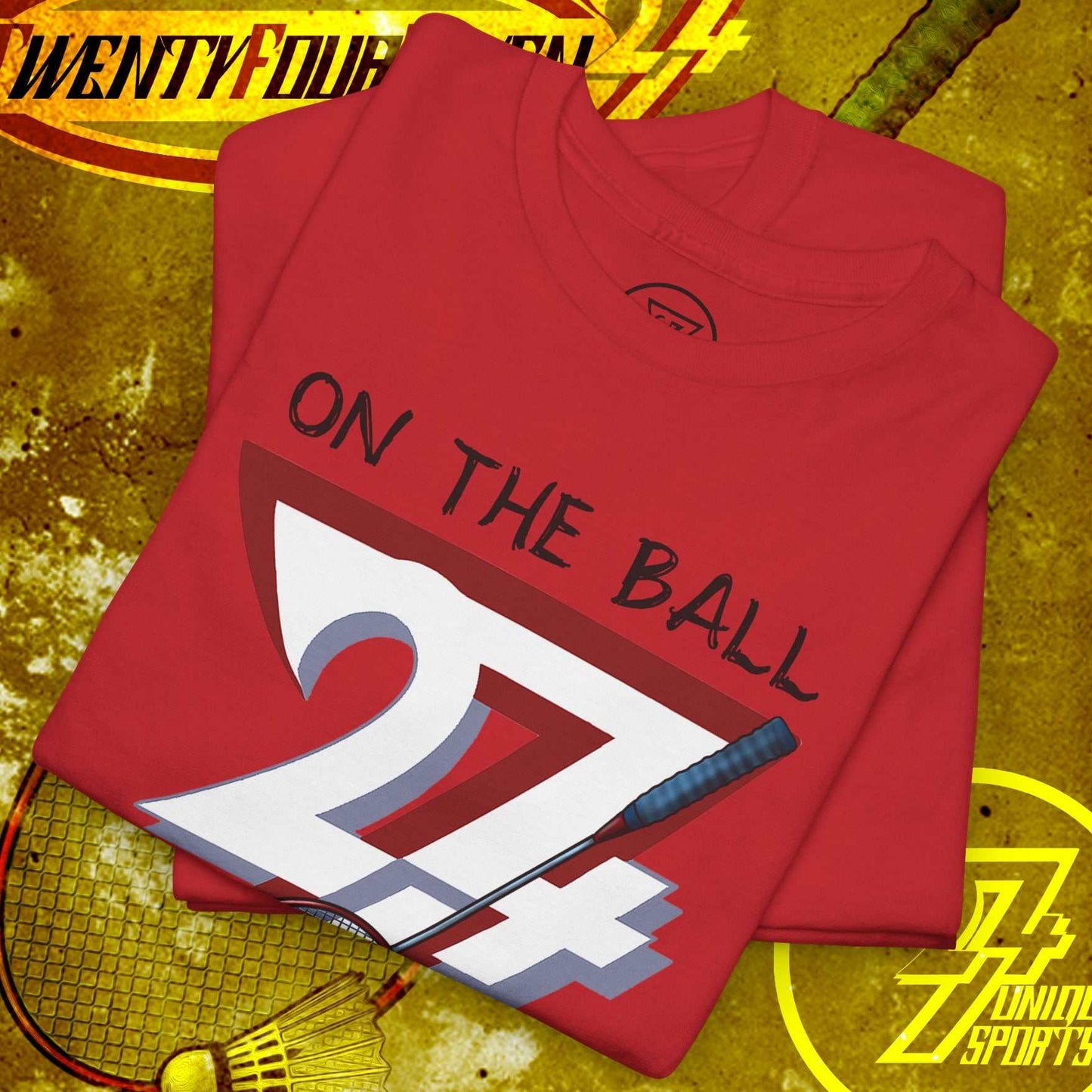 Red badminton tee with hand-drawn graphic showing "On the Ball 24/7" text, badminton racket, and shuttlecock design, laid on a textured yellow background.