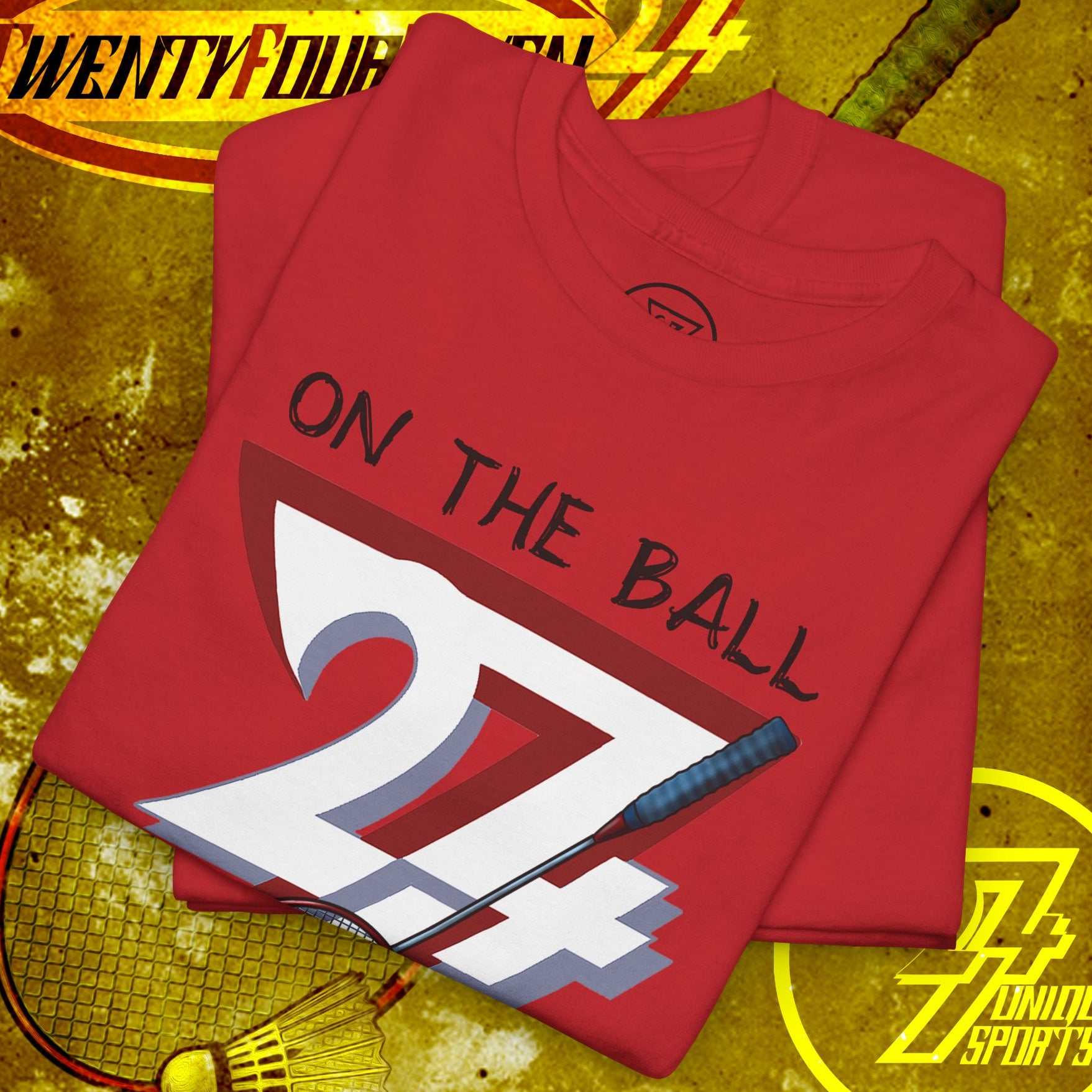 Red badminton tee with hand-drawn graphic showing "On the Ball 24/7" text, badminton racket, and shuttlecock design, laid on a textured yellow background.