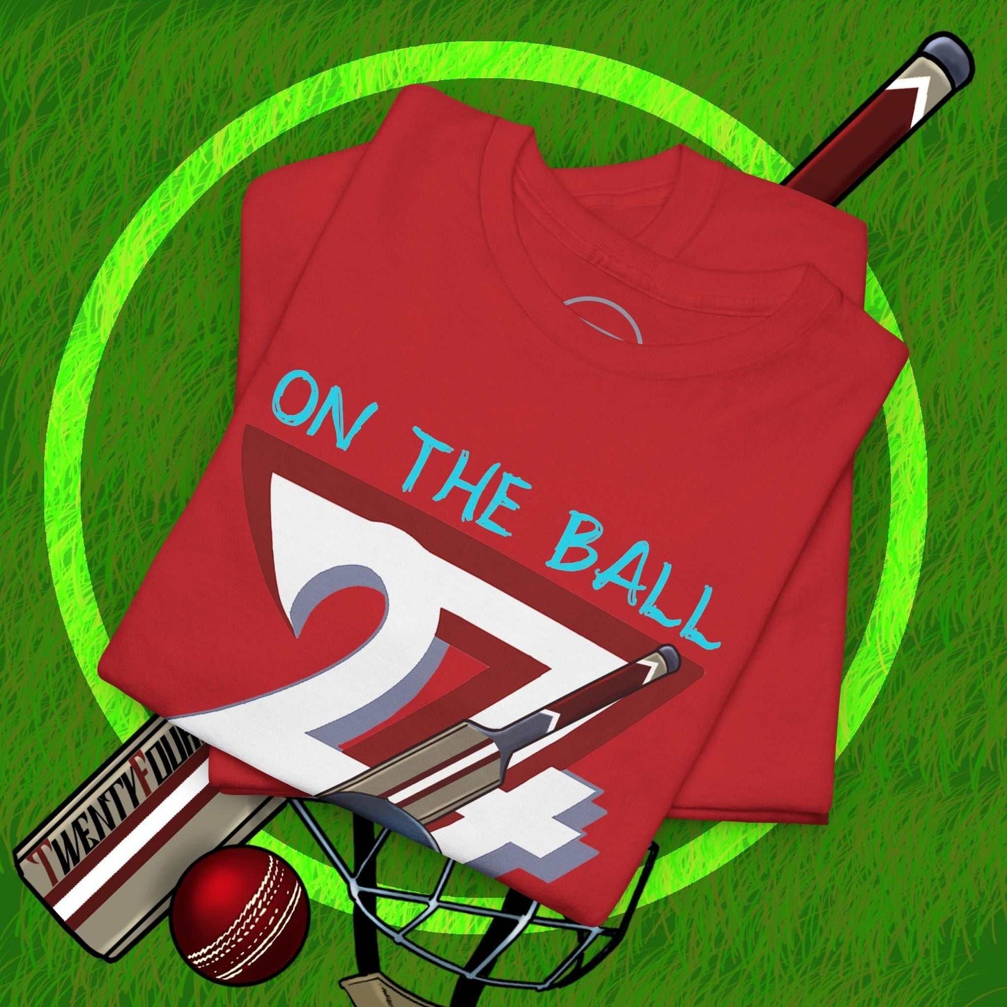 Red cricket tee with "On the Ball 24/7" hand-drawn vintage cricket design, folded on green grass with cricket bat, ball, and helmet accessories.
