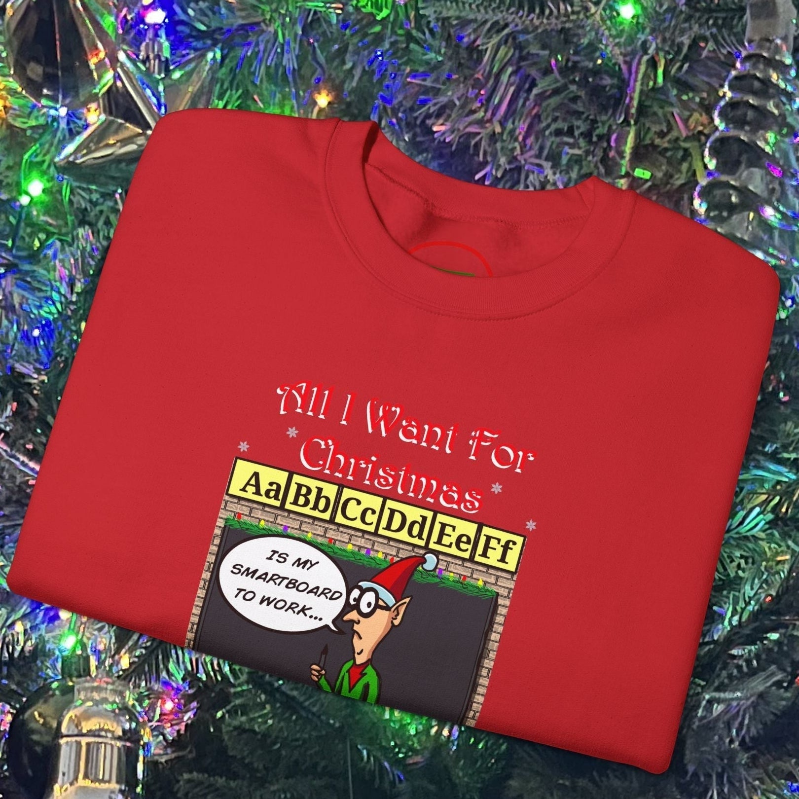 Red funny cartoon teacher Christmas sweatshirt with humorous holiday design and classroom theme, folded on Christmas tree background