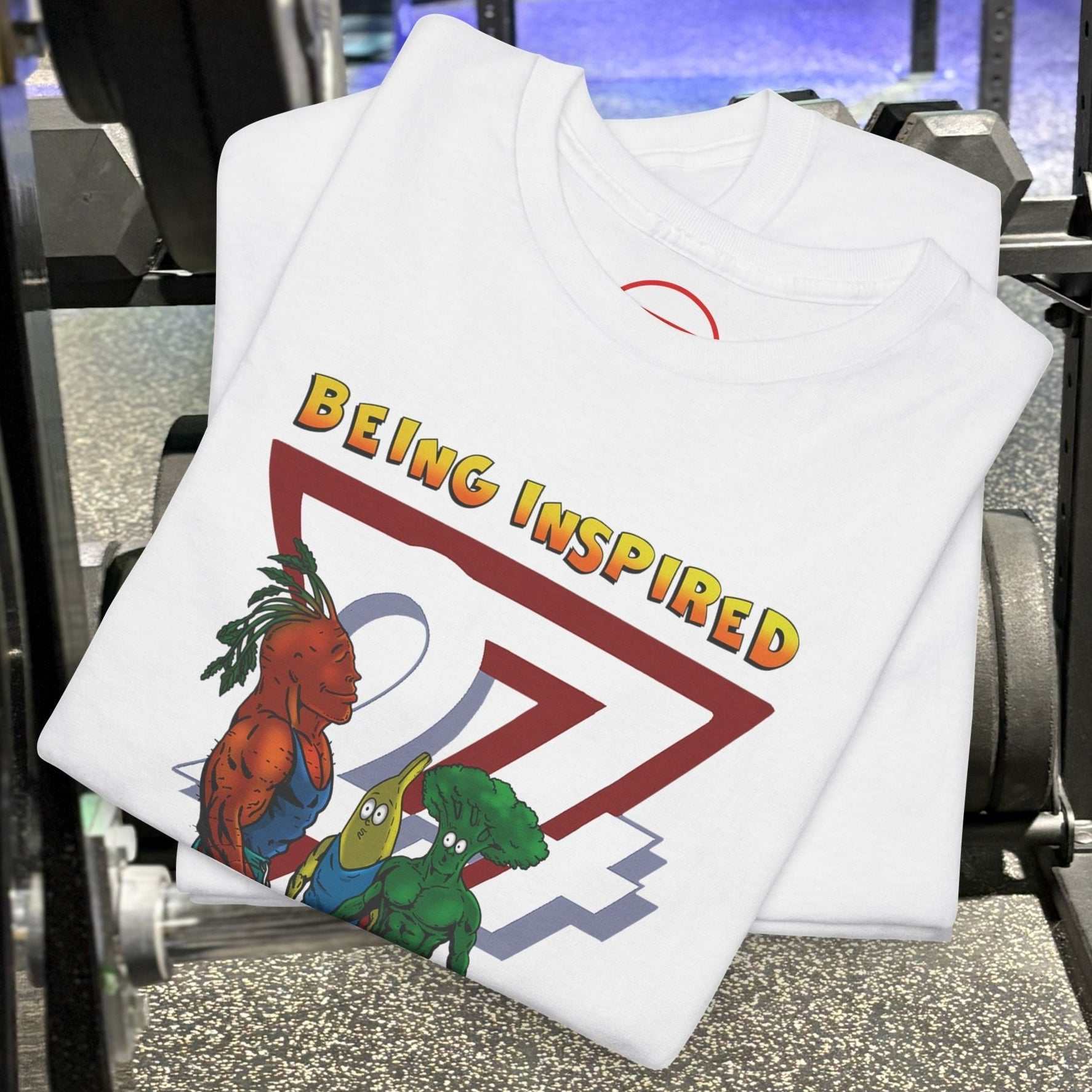 White Funny Carrot Gym Tee with cartoon carrot, banana, and broccoli lifting weights and "Being Inspired" text, folded on gym floor