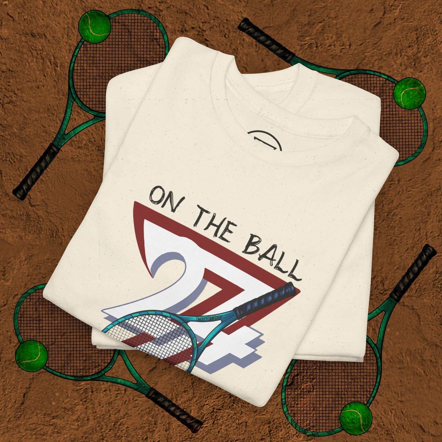 White tennis tee with hand-drawn "On the Ball 24/7" design folded on clay tennis court background with rackets and balls