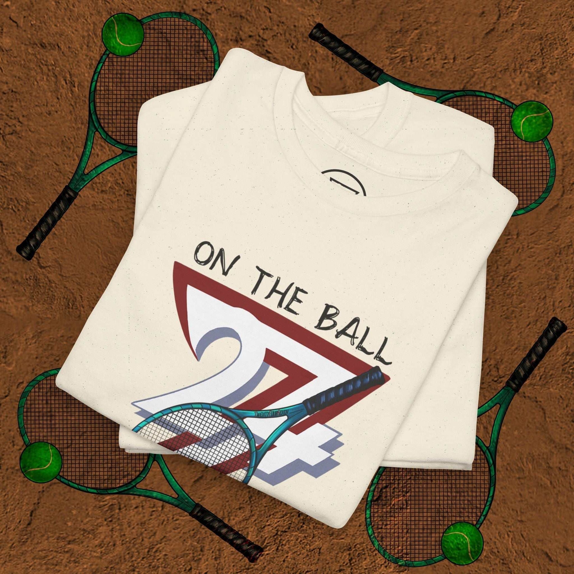 White tennis tee with hand-drawn "On the Ball 24/7" design folded on clay tennis court background with rackets and balls