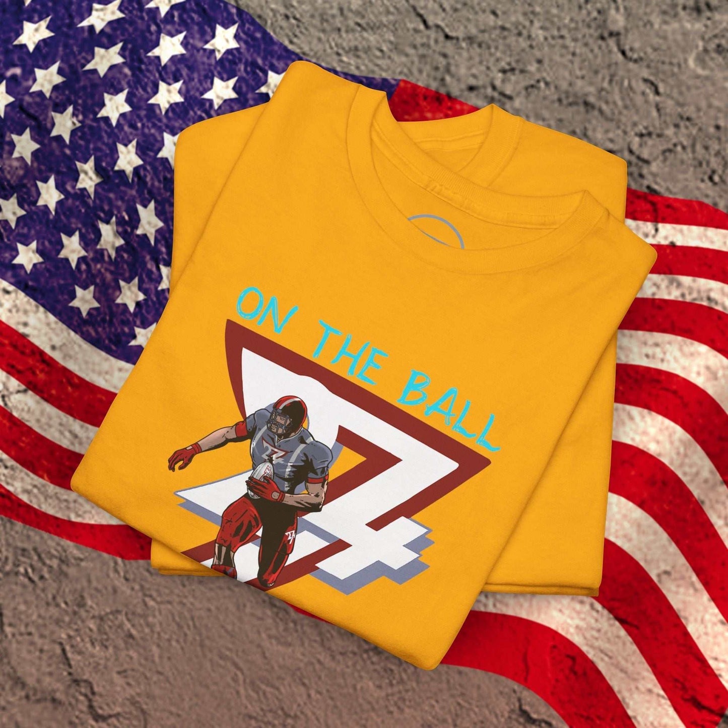 Yellow American football tee with hand-drawn comic-style player illustration and "ON THE BALL" text, folded on American flag background
