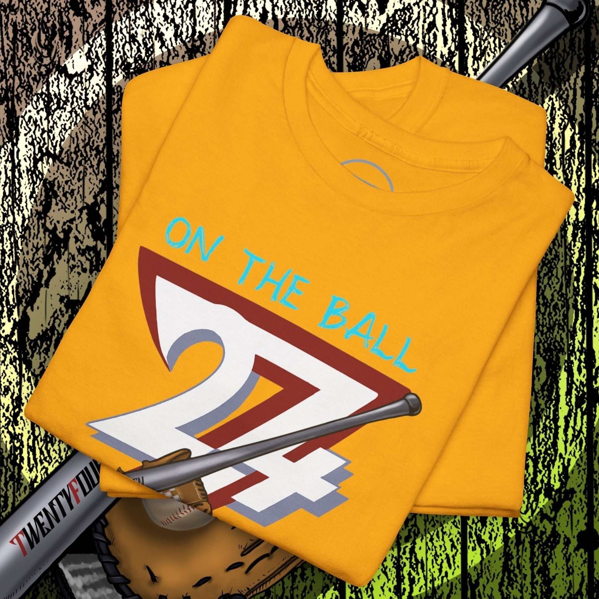 Yellow baseball illustrated tee with hand-drawn "On the Ball 24/7" design, folded on baseball glove and bat background