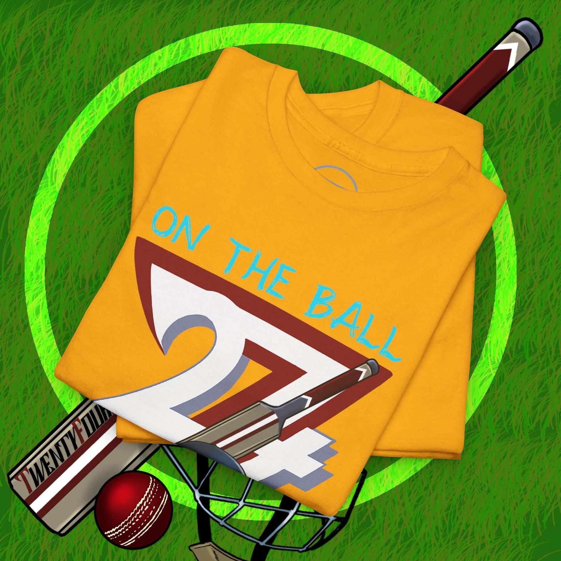 Yellow cricket tee with hand-drawn 90s style "On The Ball" illustration, folded on grass with cricket bat, ball, and helmet