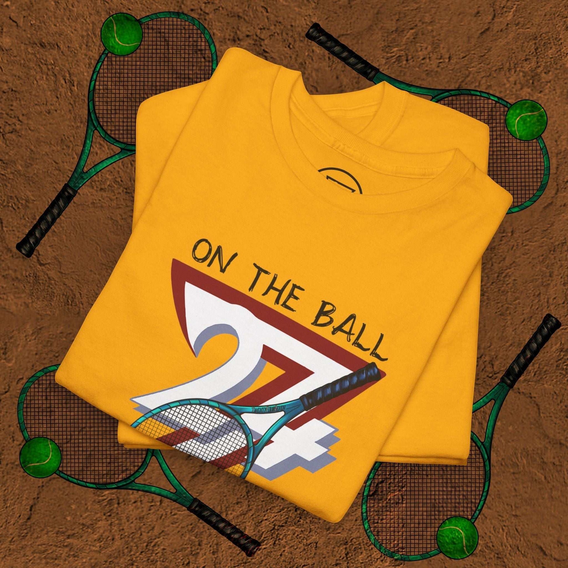 Yellow tennis tee with "On the Ball 24/7" hand-drawn sports design, folded on a textured brown surface surrounded by tennis rackets and balls
