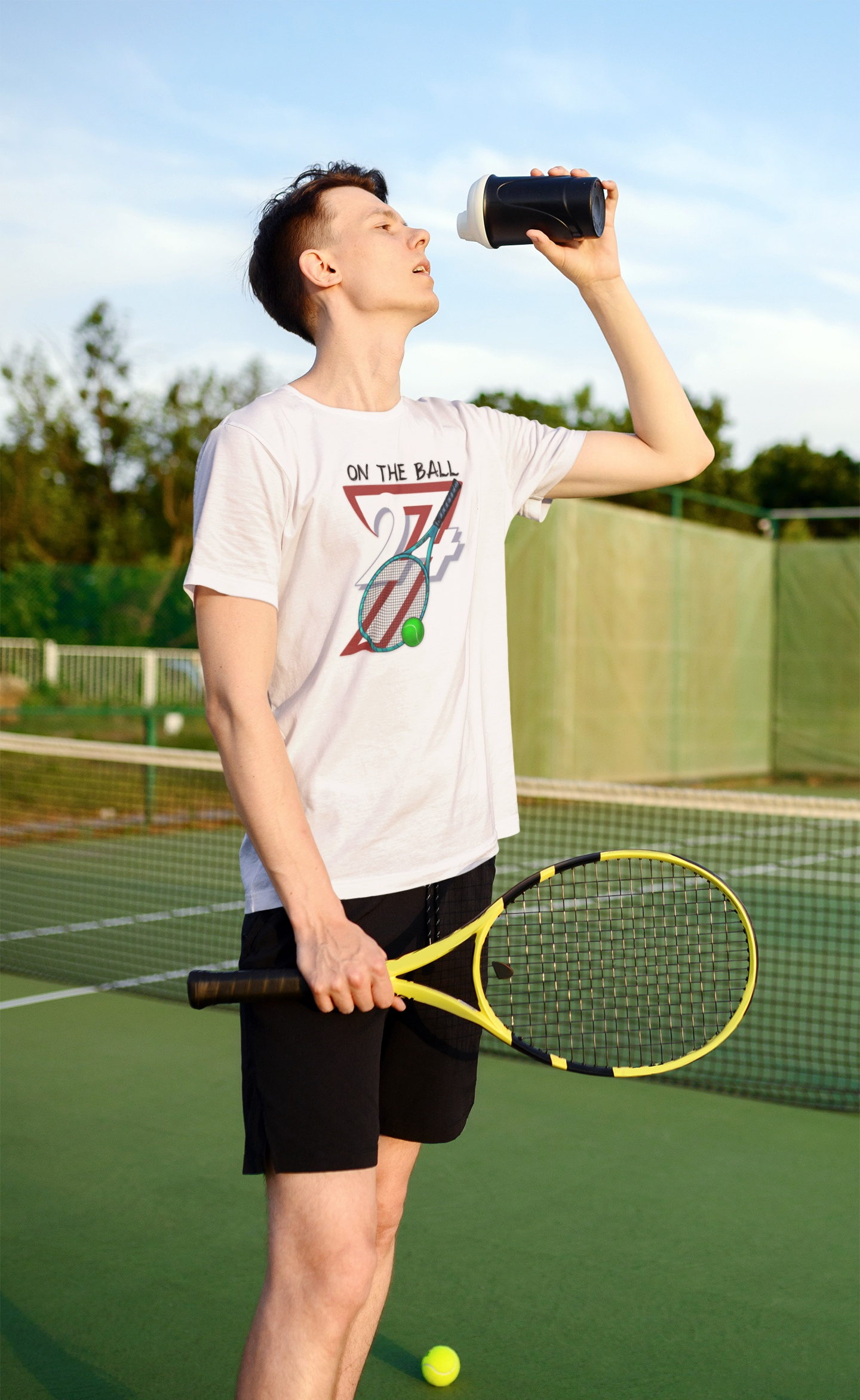 Young man wearing tennis tee with "On the Ball 24/7" design holding tennis racket and drinking water on outdoor court