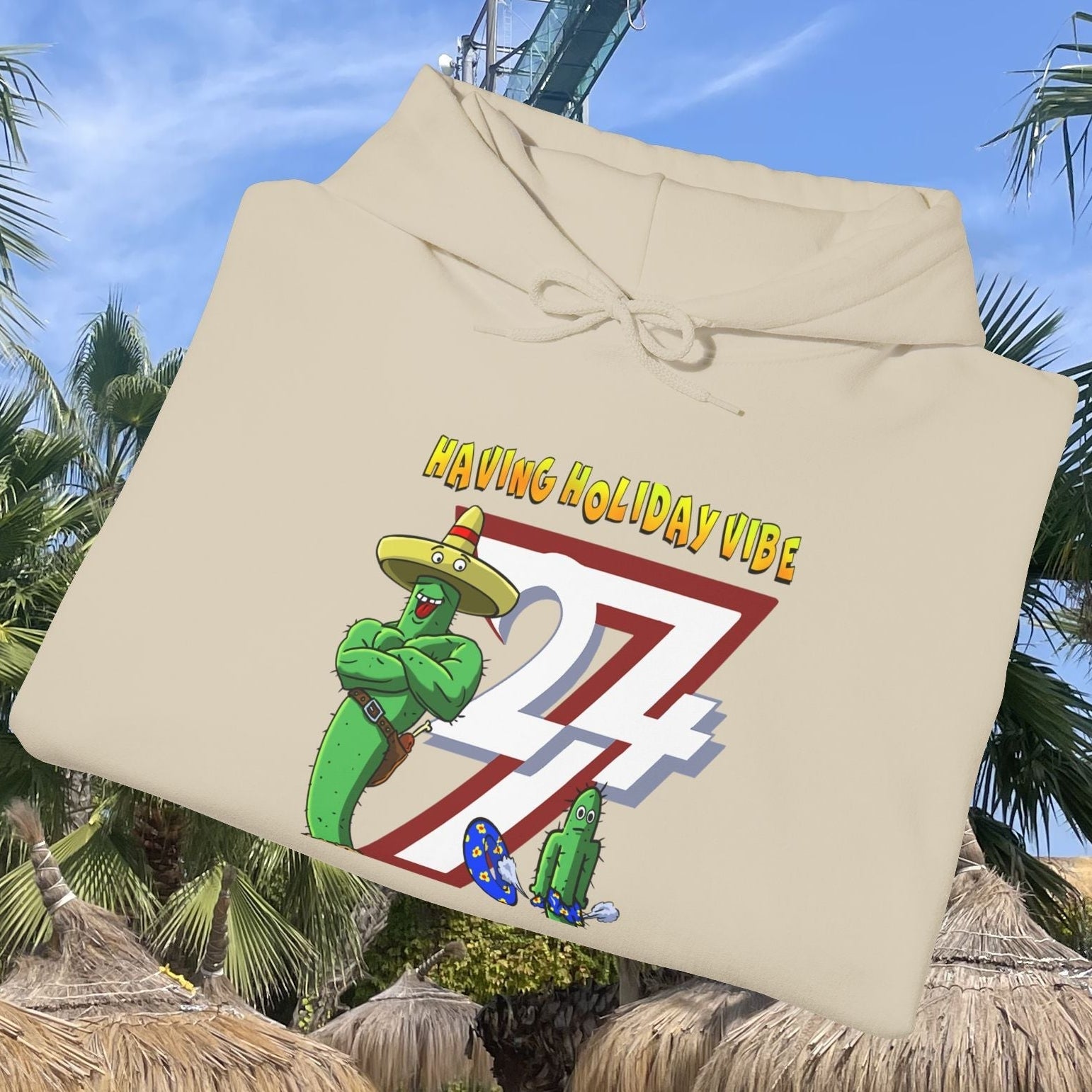 beige cactus hoodie with cartoon holiday vibe design displayed outdoors among palm trees and thatched umbrellas