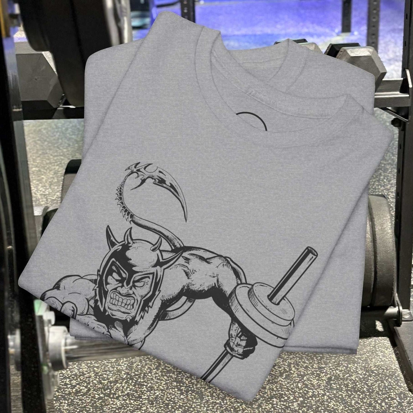 gray gym tee with comic-style muscular devil holding barbell illustration, folded on gym floor next to equipment
